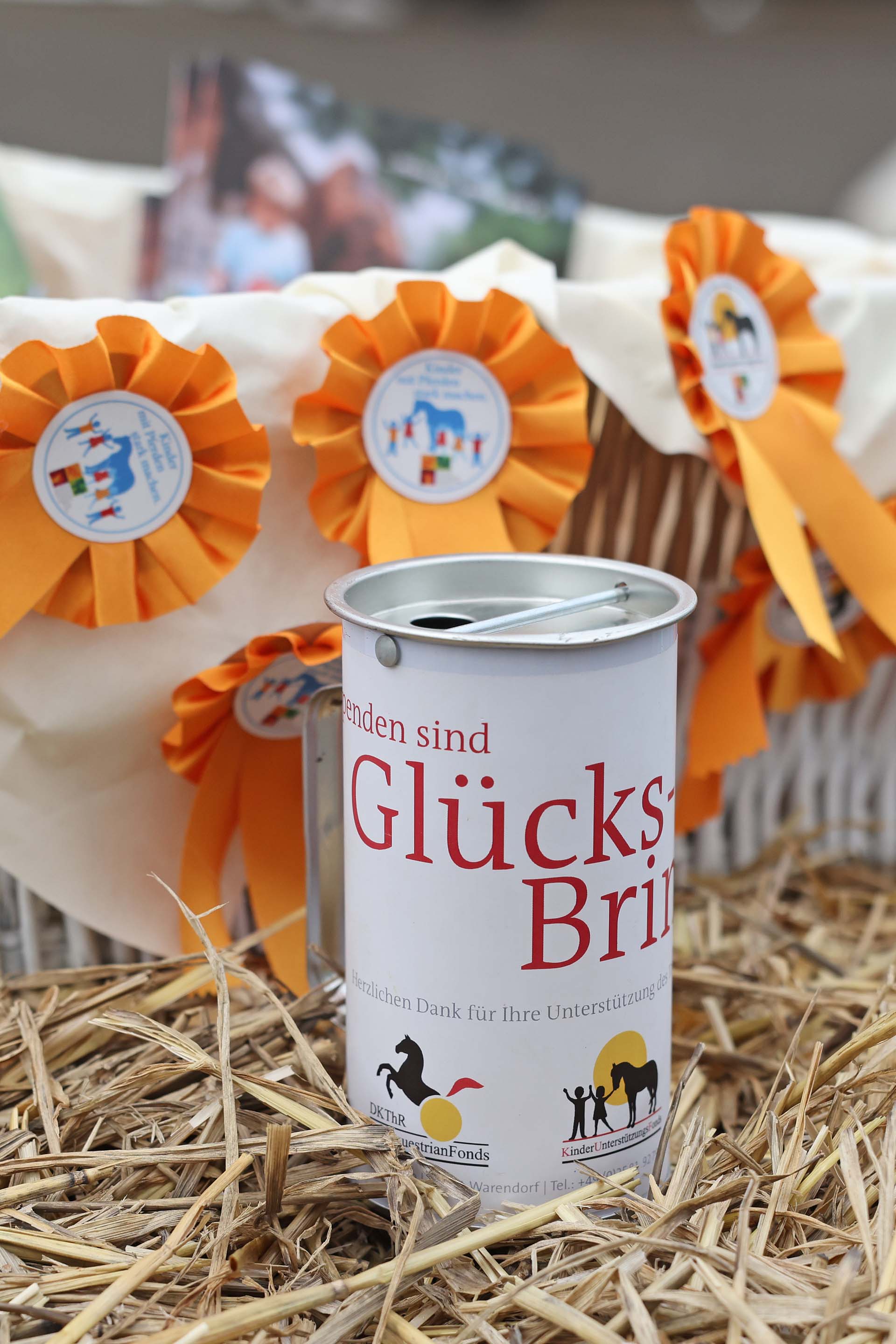 CHIO Aachen | Glücks-Bringer | ALRV - DKThR | Foto: Andreas Steindl Eine Spendendose mit der Aufschrift "Glücks-Bringer" steht auf einer Heubodenfläche, umgeben von orangefarbenen Schleifen mit Aufklebern. Im Hintergrund befinden sich leicht verschwommene Bilder von Kindern und Pferden.