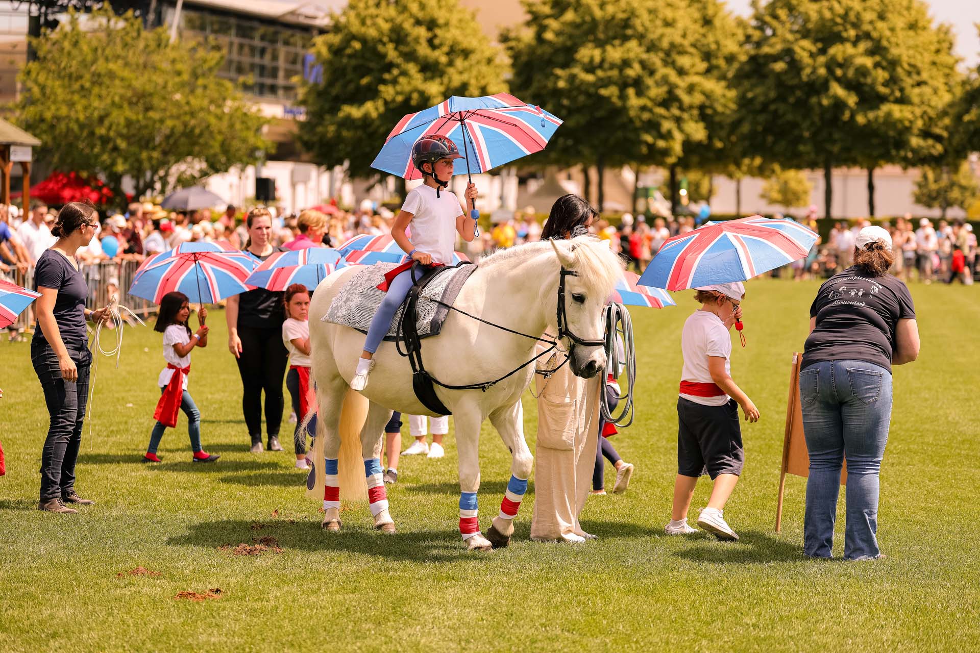 CHIO Aachen | Schaubild DKThR | Foto: Andreas Steindl Ein Kind sitzt auf einem weißen Pferd und hält einen Regenschirm mit rot-blauem Design. Um sie herum stehen mehrere Personen, darunter Kinder mit ähnlichen Regenschirmen, während eine große Menschenmenge im Hintergrund zu sehen ist. Der Schauplatz ist ein grünes Feld unter blauem Himmel.