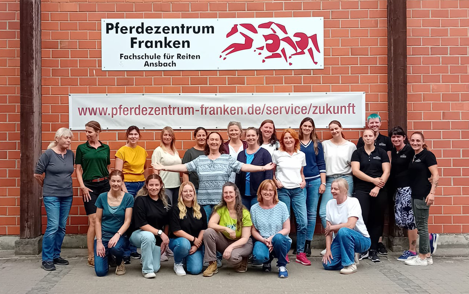 Gruppenfoto von Personen vor dem Pferdezentrum Franken in Ansbach, darunter mehrere Frauen in verschiedenen Outfits, die zusammenstehen, während ein Banner mit der Website-Adresse und dem Logo des Zentrums im Hintergrund sichtbar ist.
