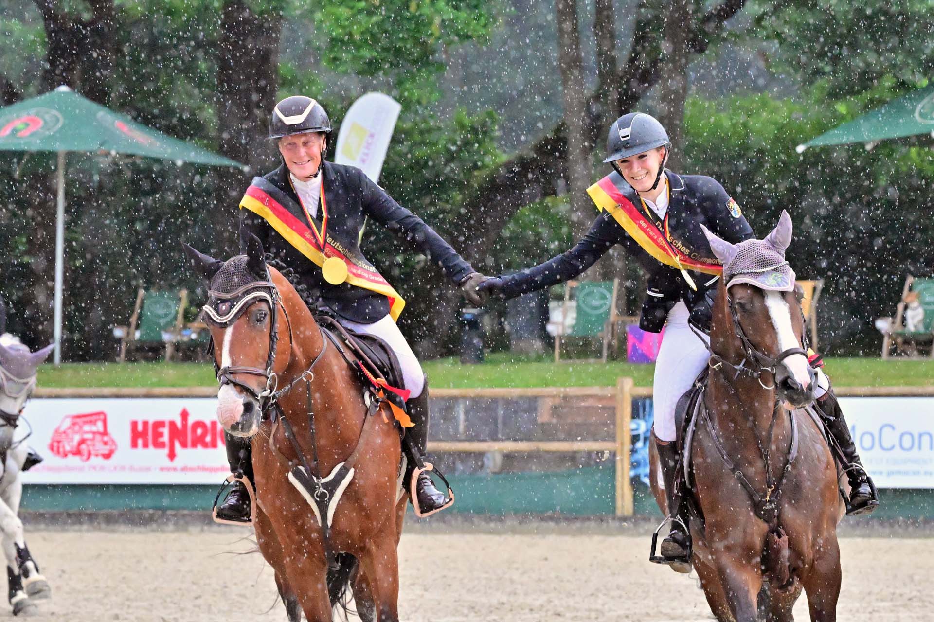 Zwei Reiterinnen in festlicher Kleidung reiten auf ihren Pferden, während sie sich in einem beeinflussten Regen umarmen. Beide tragen Medaillen und sind mit Schärpen geschmückt, die auf eine Meisterschaft hinweisen.