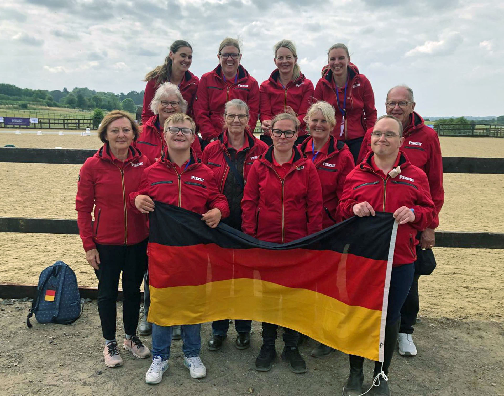 VIRTUS World Equestrian Dressage Championships in Winchester | Team & Team behind the Team, Foto: Privat Eine Gruppe von zehn Personen in roten Jacken steht vor einem Reitplatz, einige halten eine deutsche Flagge. Im Hintergrund sind Hecken und ein sandiger Boden sichtbar.