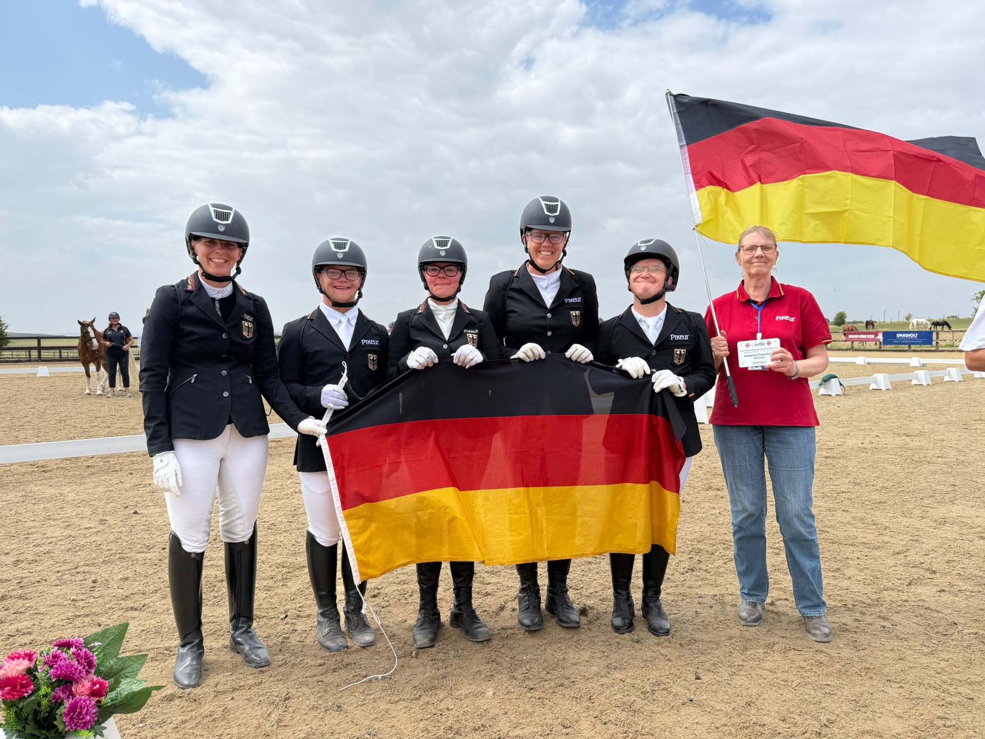 VIRTUS World Equestrian Dressage Championships in Winchester / Großbritannien| Team Deutschland | Foto: Marion Hupperts Eine Gruppe von fünf Reitern in schwarzer Turnierbekleidung und Helm posiert auf einem Reitplatz, während sie eine deutsche Flagge halten. Im Hintergrund ist ein weiteres Pferd und eine Person zu sehen. Die Umgebung ist sandig, mit einer blauen Himmel und einigen Wolken.