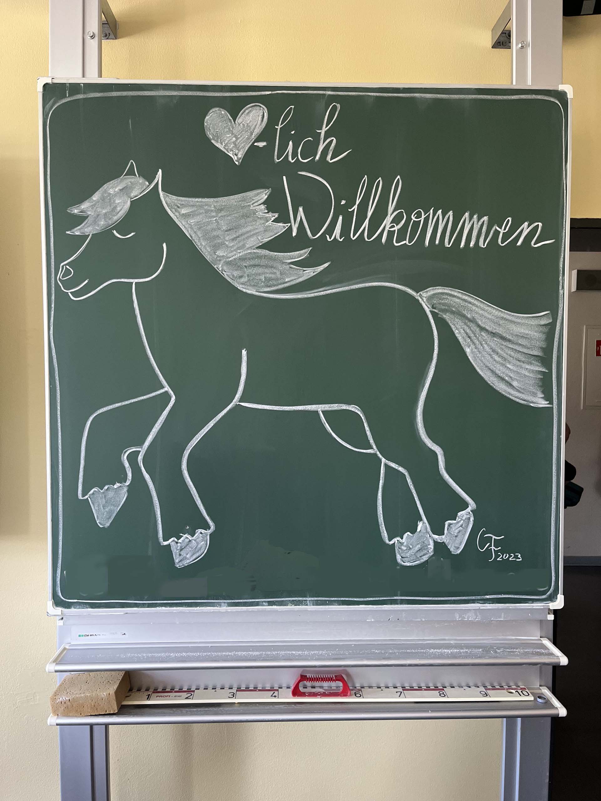 Eine grüne Tafel zeigt eine zeichnerische Darstellung eines Pferdes in Bewegung, umgeben von dem Schriftzug 'Herzlich Willkommen'.