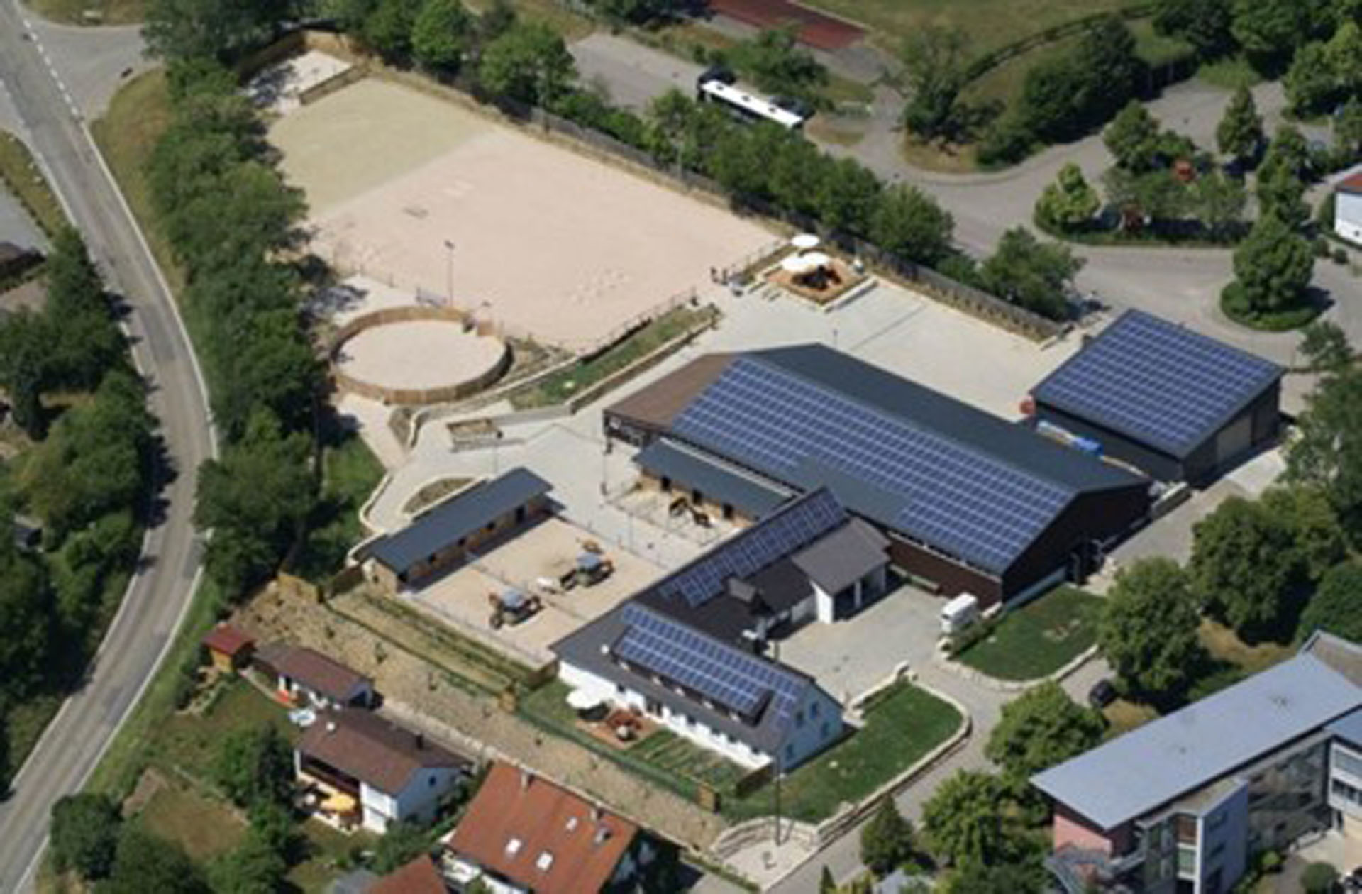 Luftaufnahme eines weitläufigen Reiterhofs mit mehreren Stallgebäuden, einem Sandplatz für das Reiten, einer rundem Pferdeplatz, umgeben von Bäumen und Wohngebäuden in der Nähe einer Straße.