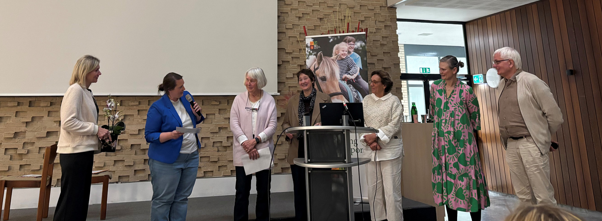Eine Gruppe von sieben Personen steht auf einer Bühne in einem modernen Veranstaltungsraum. Eine Frau mit Mikrofon spricht zu den Anwesenden, während die anderen aufmerksam zuhören. Auf einem Bildschirm im Hintergrund ist ein Bild von Kindern mit einem Pferd zu sehen. Die Frauen tragen verschiedene Kleidung, darunter ein pinkes Kleid mit grünen Mustern. Der Raum hat eine mit Stein verkleidete Wand und eine helle, freundliche Atmosphäre.