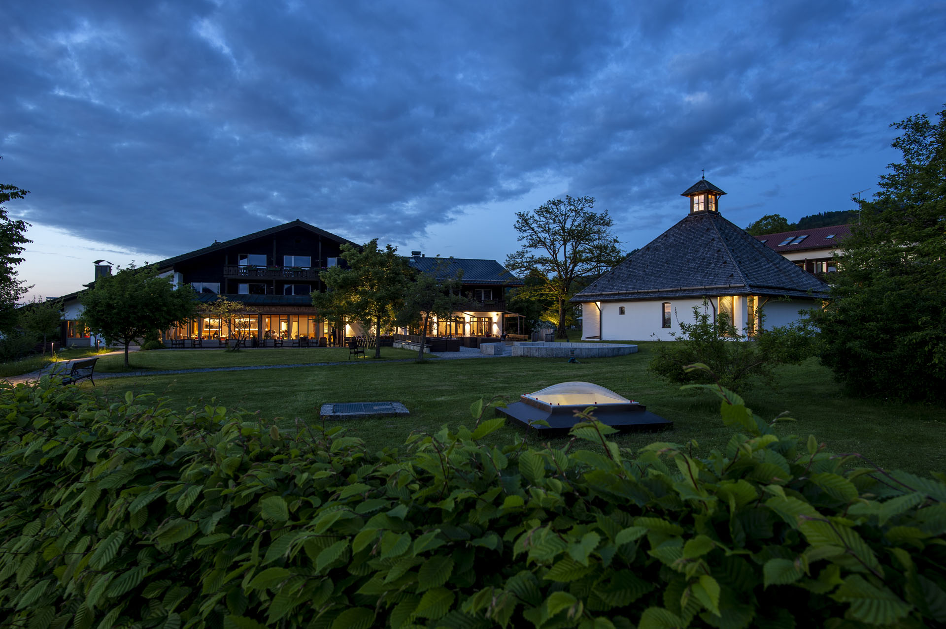 Die Außenansicht eines Hotels bei Dämmerung, präsentiert ein mehrstöckiges Gebäude mit großen Fenstern und einer angrenzenden freien Fläche, die von Bäumen und Laternen umgeben ist. In der Nähe steht ein runder, hell erleuchteter Pavillon mit einem geschindelten Dach, während im Vordergrund eine grüne Hecke zu sehen ist.