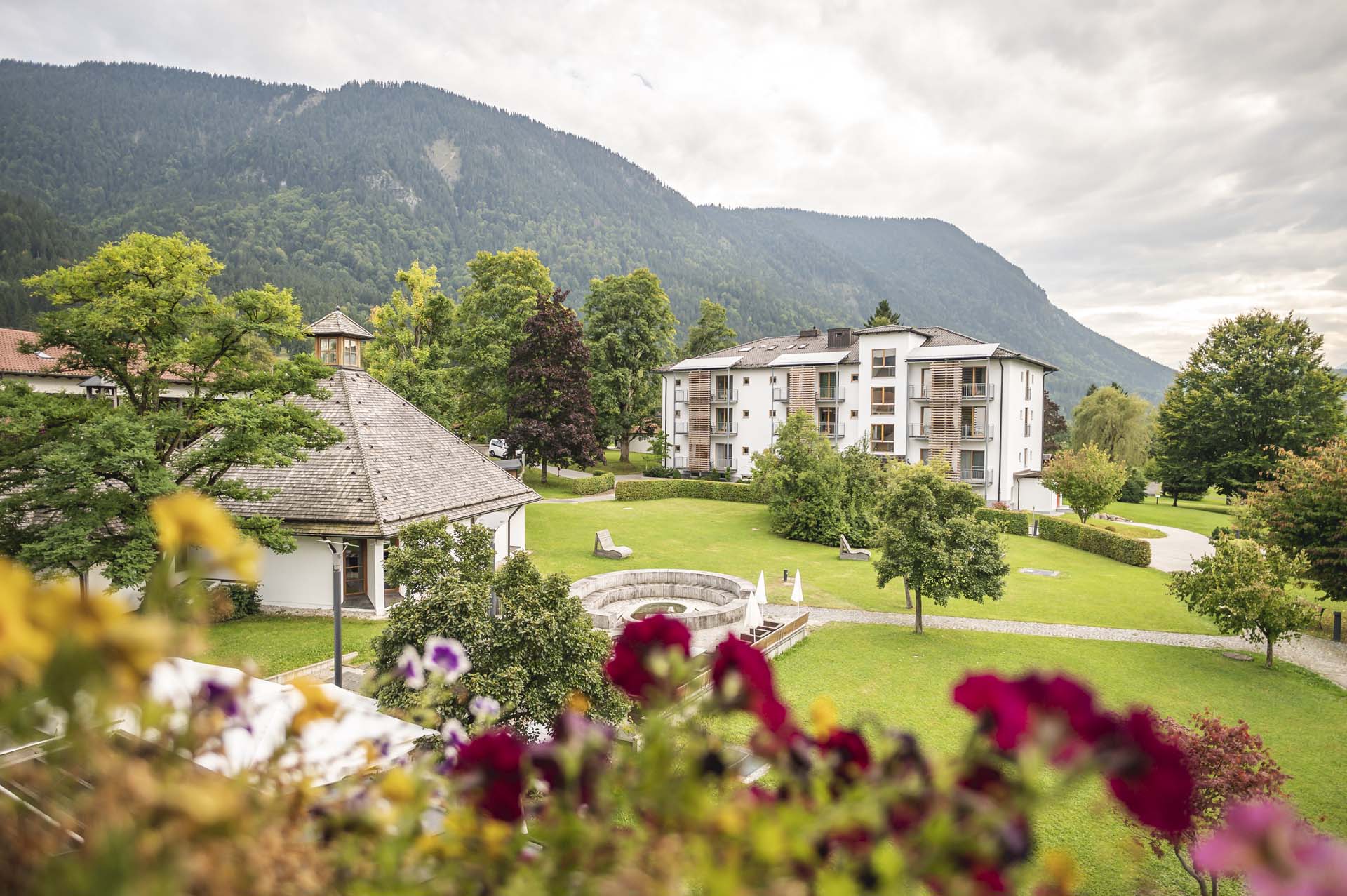 Blick auf eine grüne Gartenlandschaft mit einem modernen Gebäude im Hintergrund und einem traditionellen Pavillon im Vordergrund, umgeben von verschiedenen Bäumen und Blumen, mit Bergen im Hintergrund und einem bewölkten Himmel.