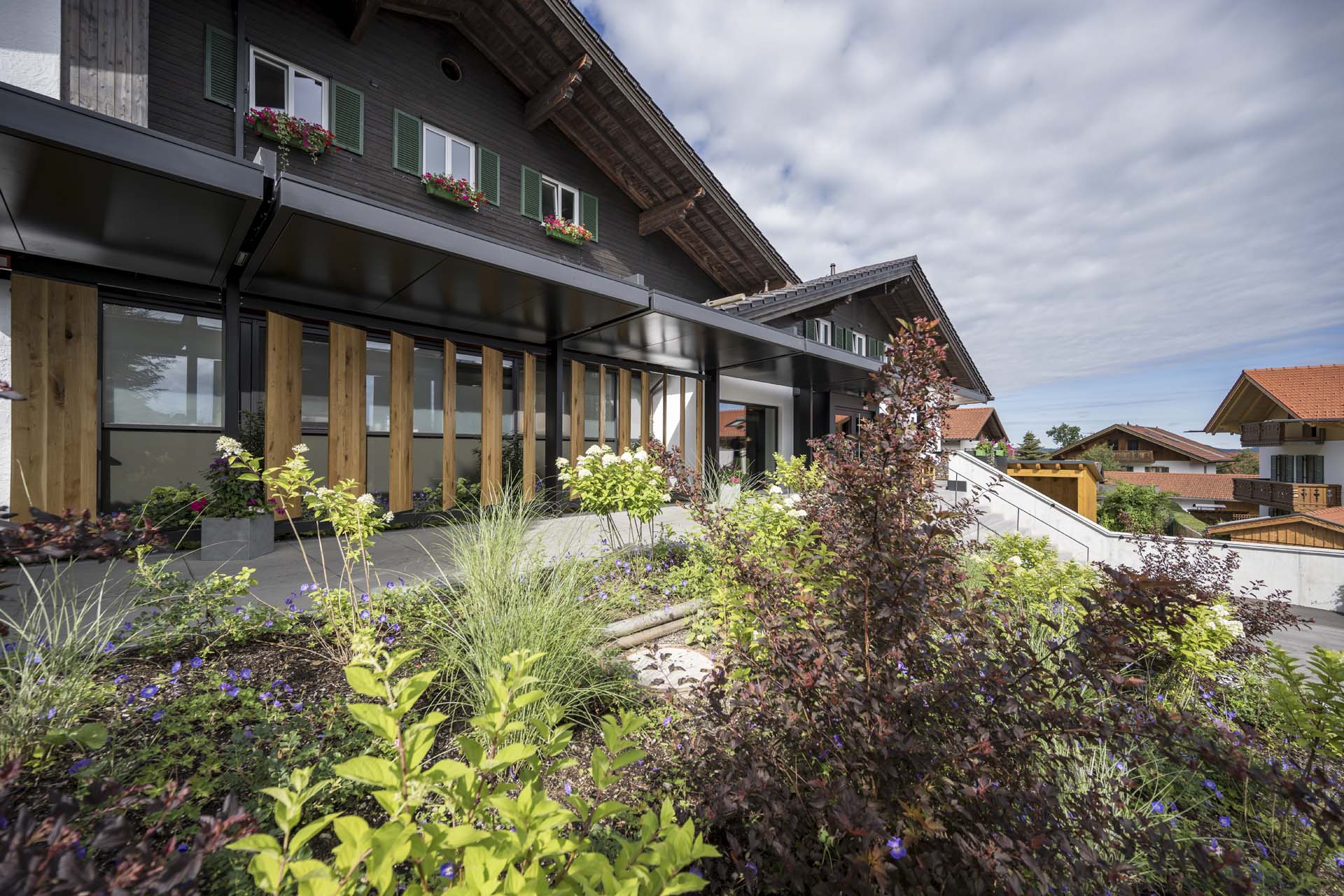 Moderne Architektur eines zweistöckigen Gebäudes mit Holz- und Glassicherungen, umgeben von üppigem Garten mit bunten Pflanzen, darunter Sträucher und Blumen, und einem klaren blauen Himmel im Hintergrund.