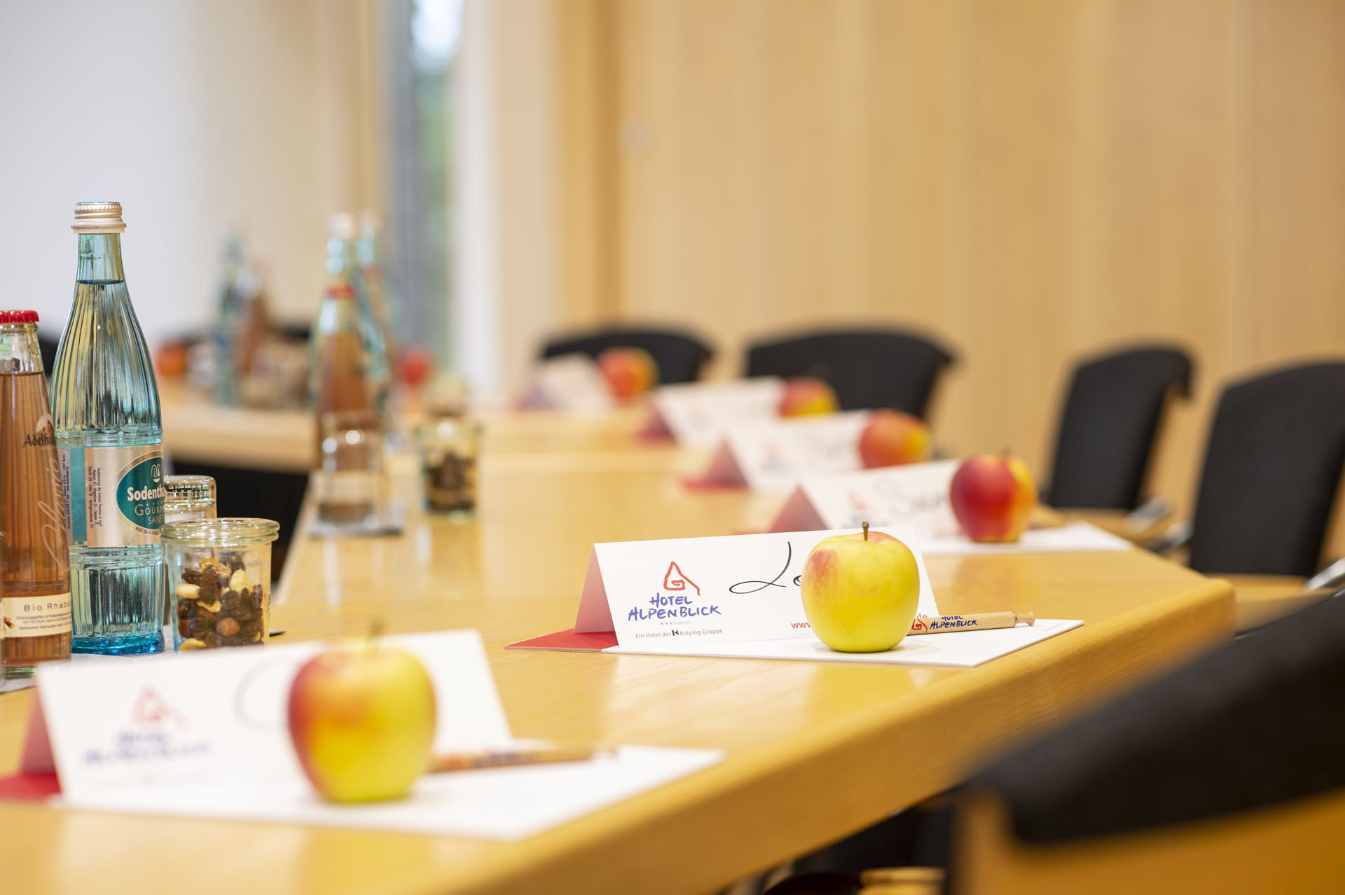 Ein Konferenztisch mit mehreren Stühlen, auf dem sich Flaschen Wasser, kleine Gläser mit Snacks und rote sowie gelbe Äpfel befinden. An jedem Platz liegt eine Karte mit dem Namen des Teilnehmers und dem Logo des Hotels Alpenblick.