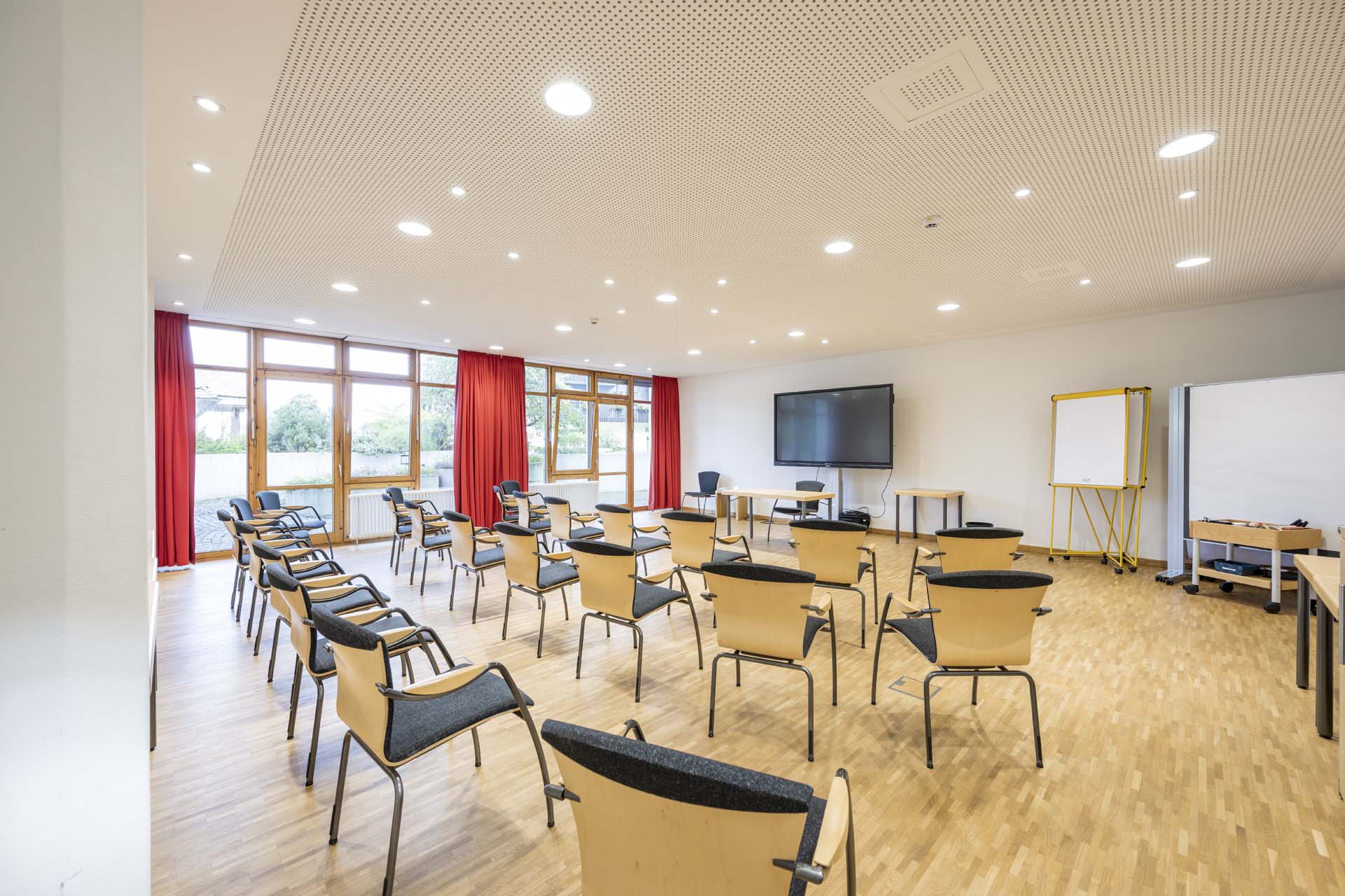 Ein heller Seminarraum mit Holzfußboden, ausgestattet mit mehreren schwarzen und hölzernen Stühlen, die in Reihen aufgestellt sind. Im Hintergrund stehen ein großer Bildschirm, ein Flipchart und ein Tisch. Rote Vorhänge rahmen die Fenster, durch die Tageslicht strömt.