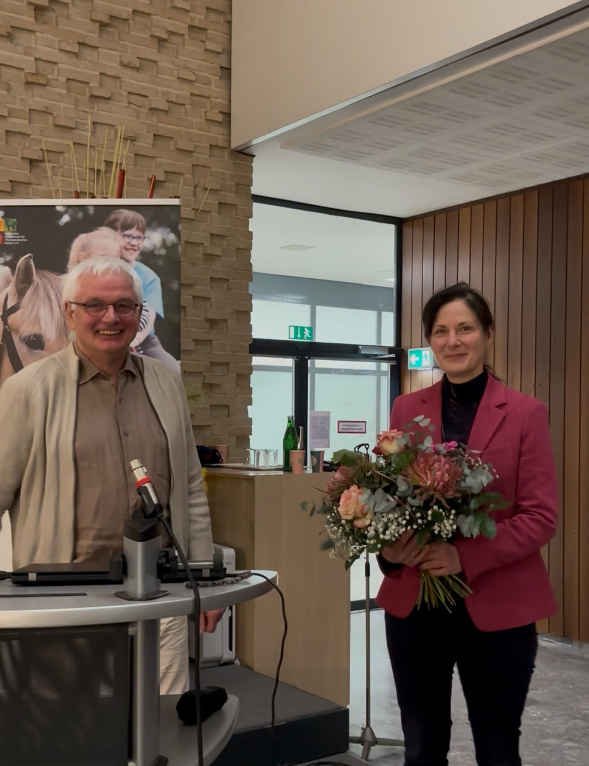 Ein Mann mit Brille und einem hellen Hemd steht neben einer Frau, die einen Blumenstrauß hält. Sie trägt einen pinken Blazer und steht an einem Rednerpult in einem modernen Raum mit Holz- und Steinwänden im Hintergrund. Im Hintergrund sind ein paar Flaschen und Getränke sichtbar.