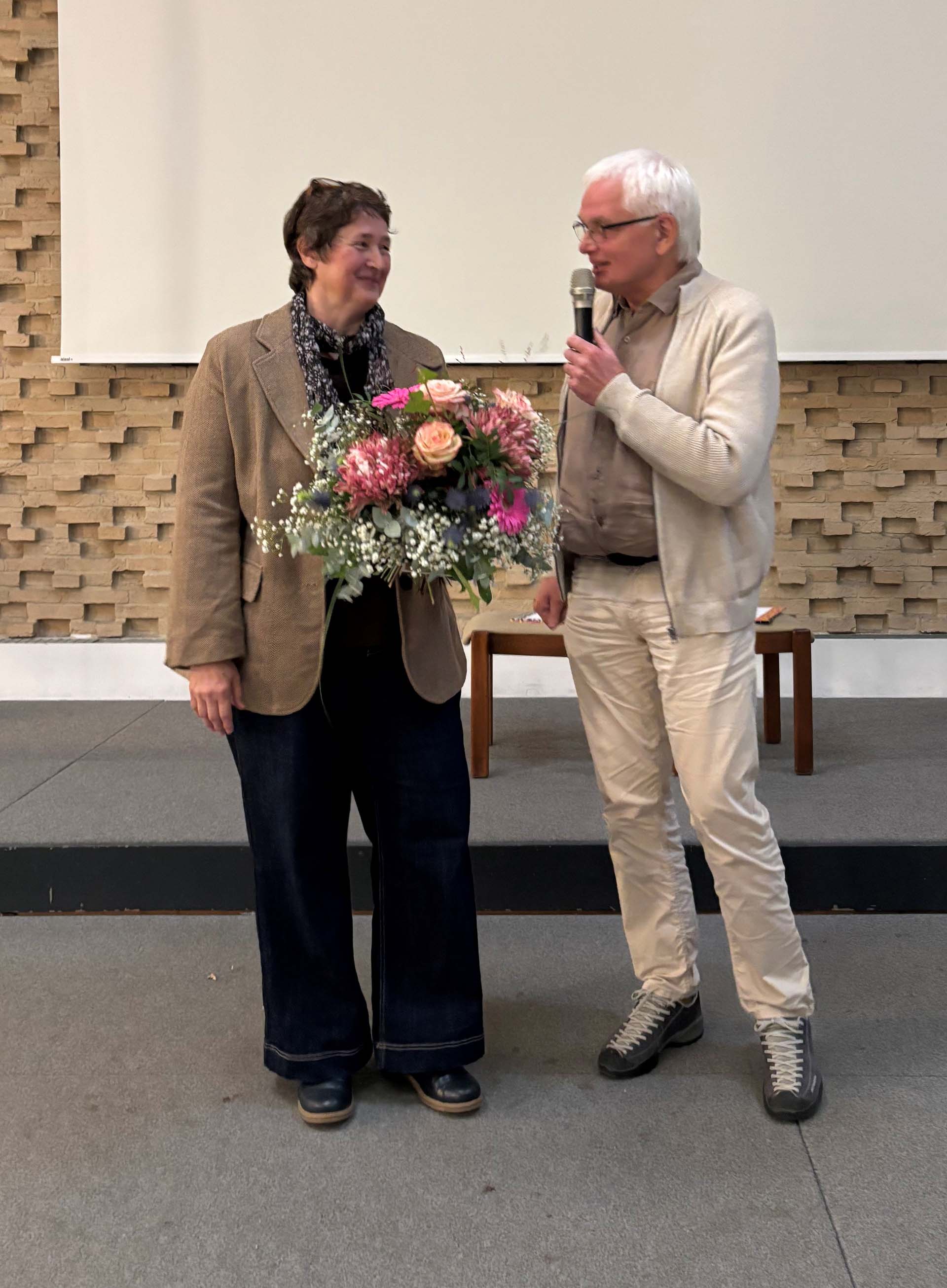 Eine Frau mit kurzen, lockigen Haaren und einer Bluse mit einem Schal steht neben einem Mann mit weißen Haaren, der ein Mikrofon hält. Die Frau hält einen großen Blumenstrauß mit rosa und lila Blumen. Im Hintergrund ist eine Wände aus Steinen und ein Tisch zu sehen.