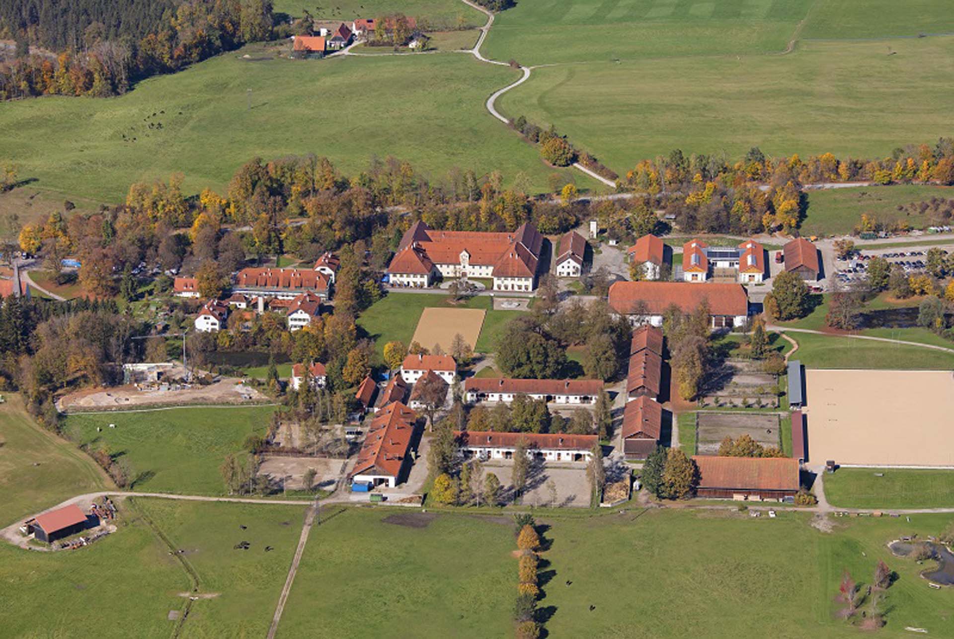 Luftaufnahme eines ländlichen Anwesens mit mehreren Gebäuden, darunter Bauernhäuser und Stallungen, umgeben von grünen Wiesen und Bäumen im Herbst mit farbigen Blättern.