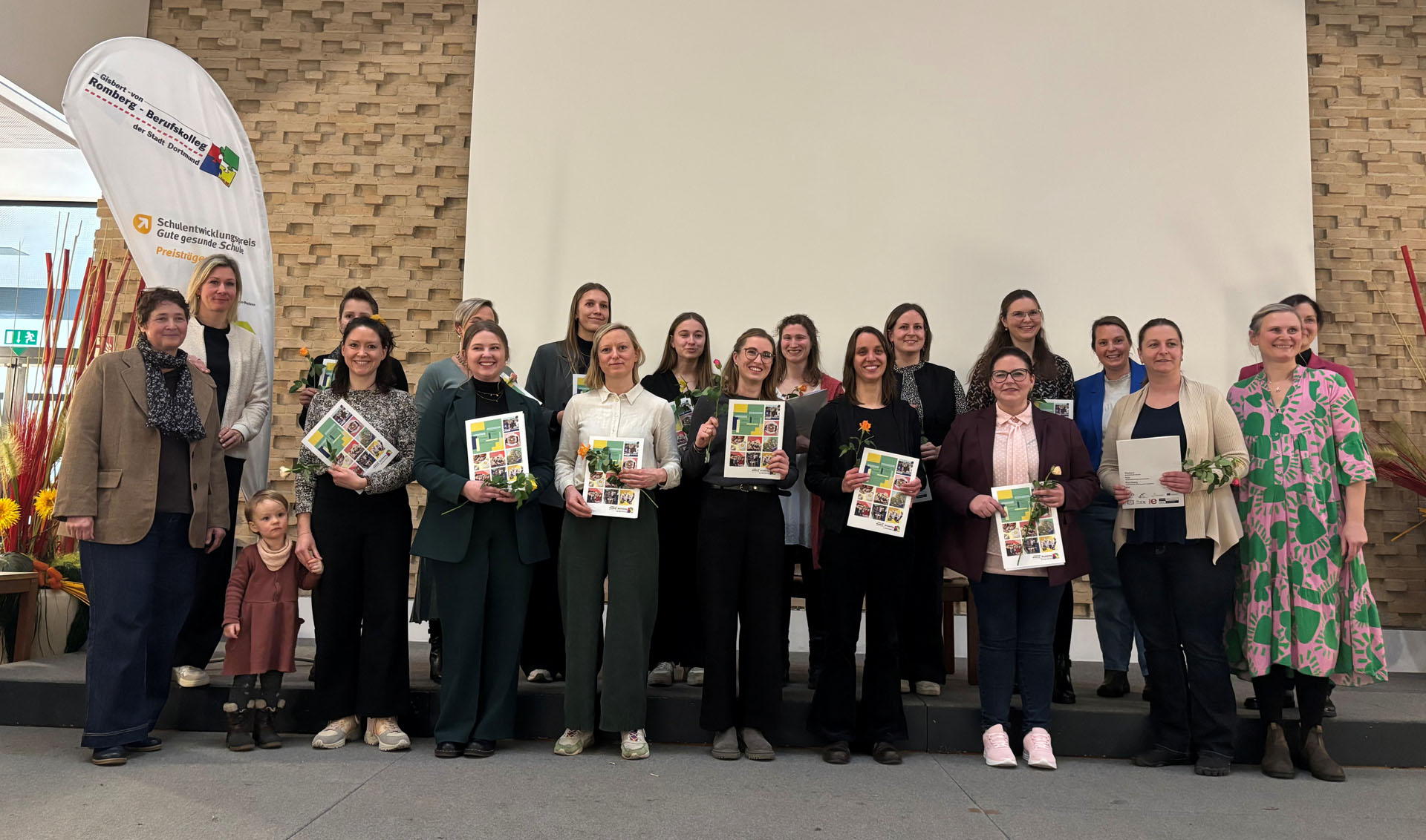Gruppenfoto von Teilnehmerinnen einer Preisverleihung, die in eleganter Kleidung stehen und Urkunden halten, während eine kleine Kind neben einer Frau steht. Im Hintergrund verweist eine Flagge auf eine Bildungseinrichtung und eine festliche Dekoration ist sichtbar.