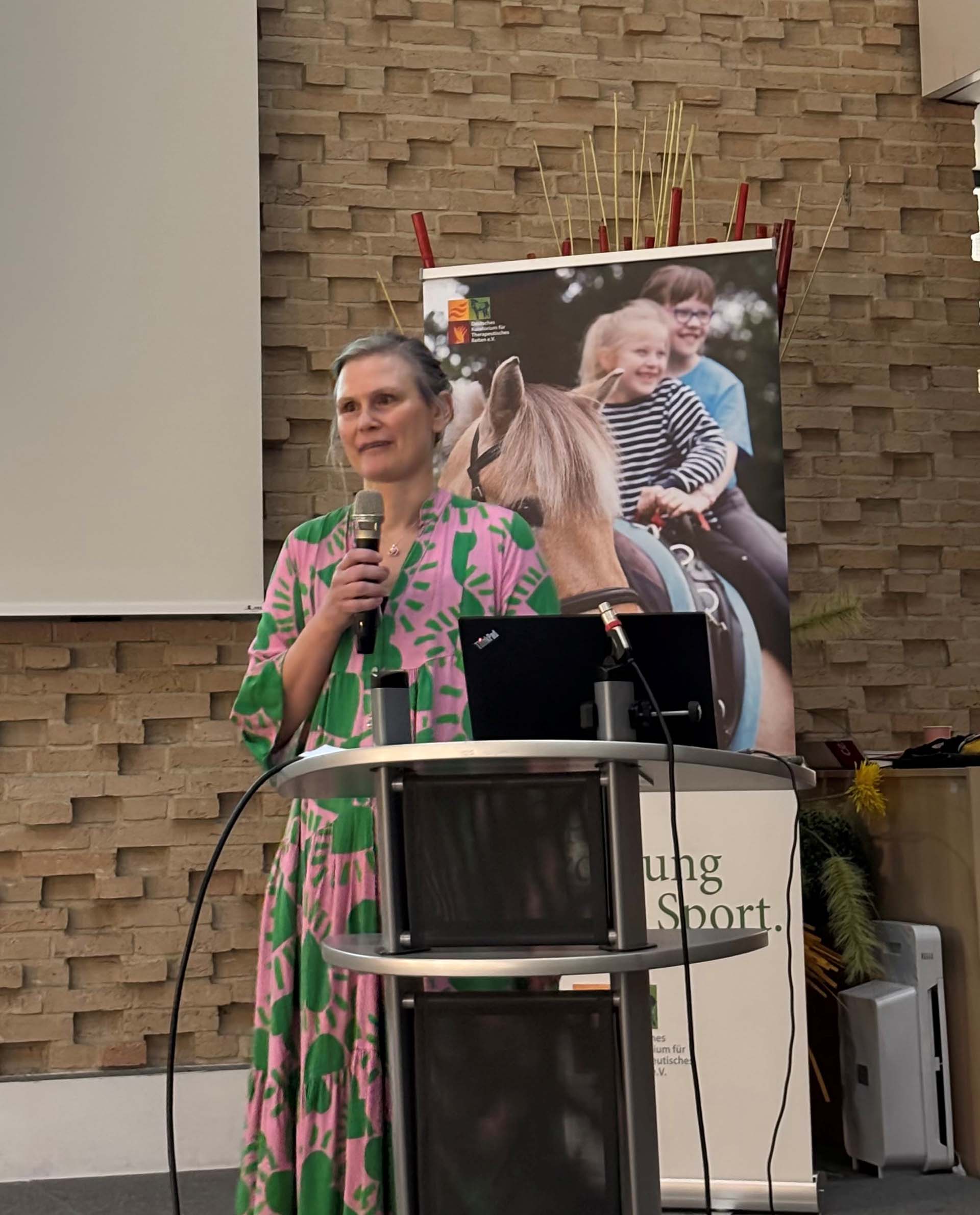 Eine Frau mit langen, blonden Haaren, die ein buntes Kleid mit grünen Mustern trägt, hält ein Mikrofon und spricht am Rednerpult in einem hellen Raum. Im Hintergrund ist ein Plakat mit lächelnden Kindern, die auf Pferden sitzen, sichtbar. An der Wand sind Steinmuster und dekorative Pflanzen angeordnet.