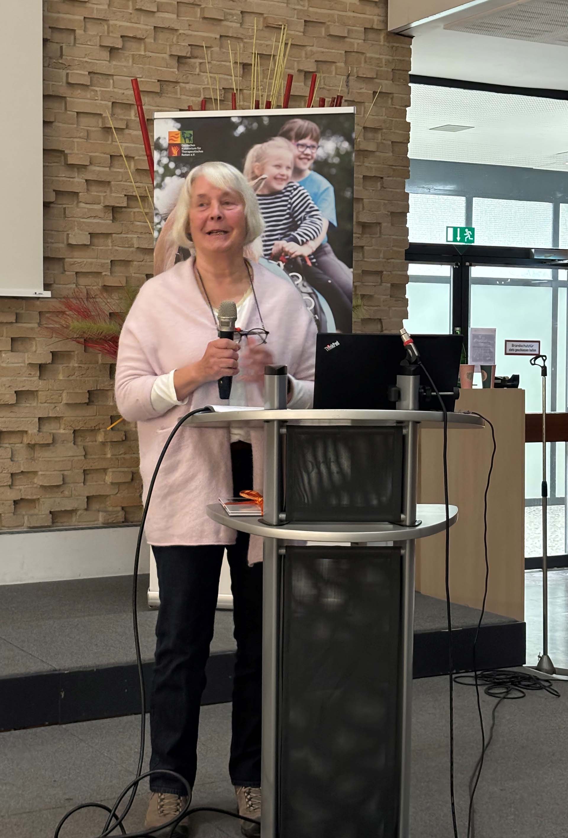 Eine Frau mit grauen Haaren, die einen rosa Pullover trägt, spricht mit einem Mikrofon in der Hand. Sie steht an einem Stehpult, während im Hintergrund ein Banner mit Kindern auf einem Fahrrad zu sehen ist. Der Raum hat eine moderne Gestaltung mit einem Sichtfenster und einer gemütlichen Atmosphäre.