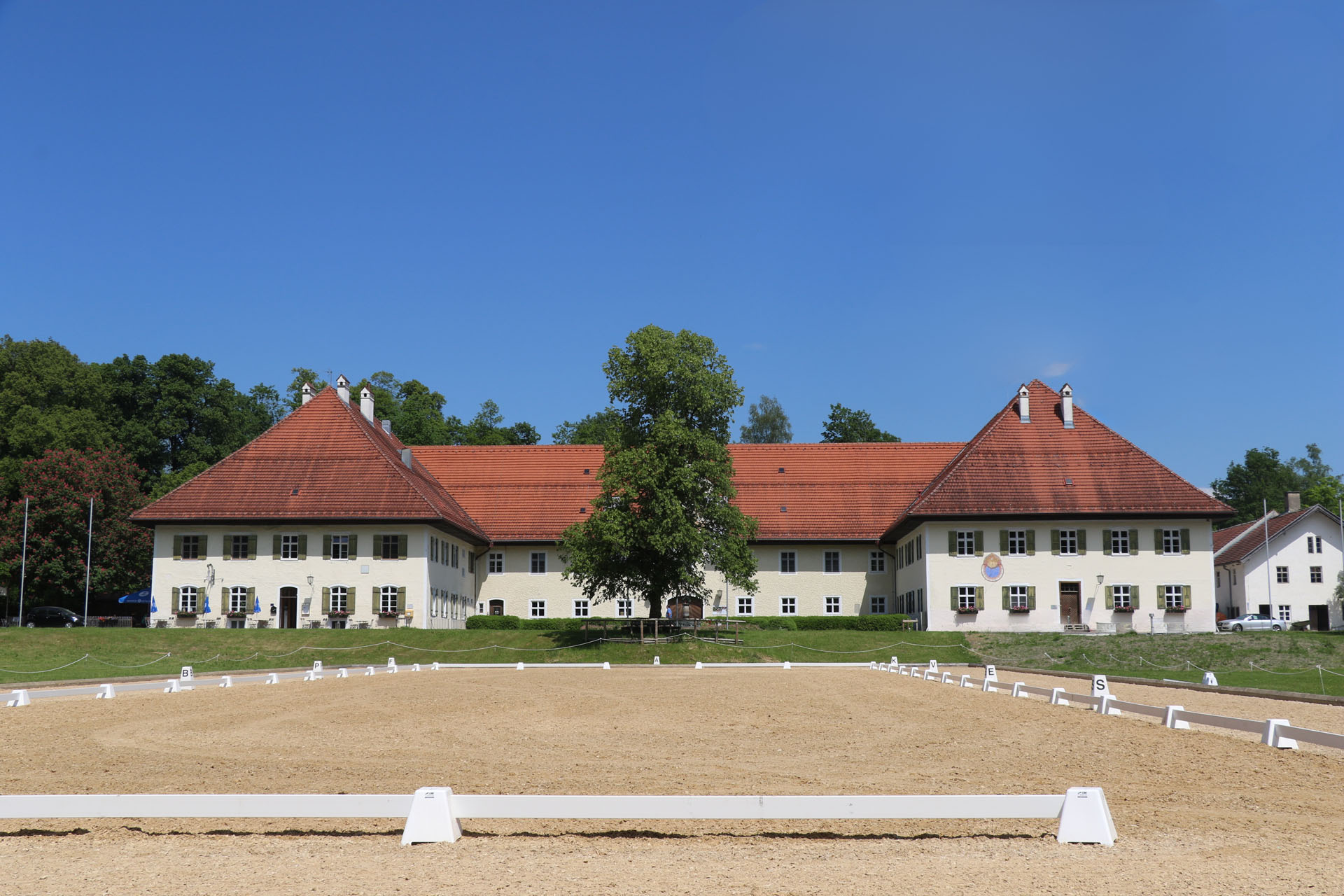 Ein großes, helles Gebäude mit hoher, roter Ziegeldachkonstruktion, umgeben von einem Pferdesportplatz mit Sandboden und weißen Markierungen, vor einem blauen Himmel und umgeben von Bäumen im Hintergrund.