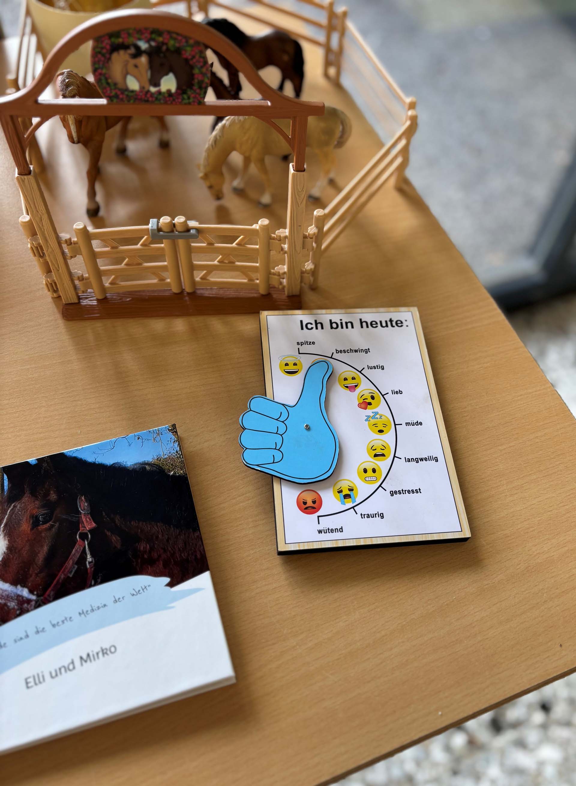 Ein Spielzeugpferch mit mehreren Kunststoffpferden, daneben liegen zwei Bücher. Eines davon zeigt ein farbenfrohes Plakat mit dem Titel 'Ich bin heute:' und verschiedenen Emojis, die unterschiedlicheEmotionen ausdrücken, sowie eine große blaue Hand mit Daumen hoch.