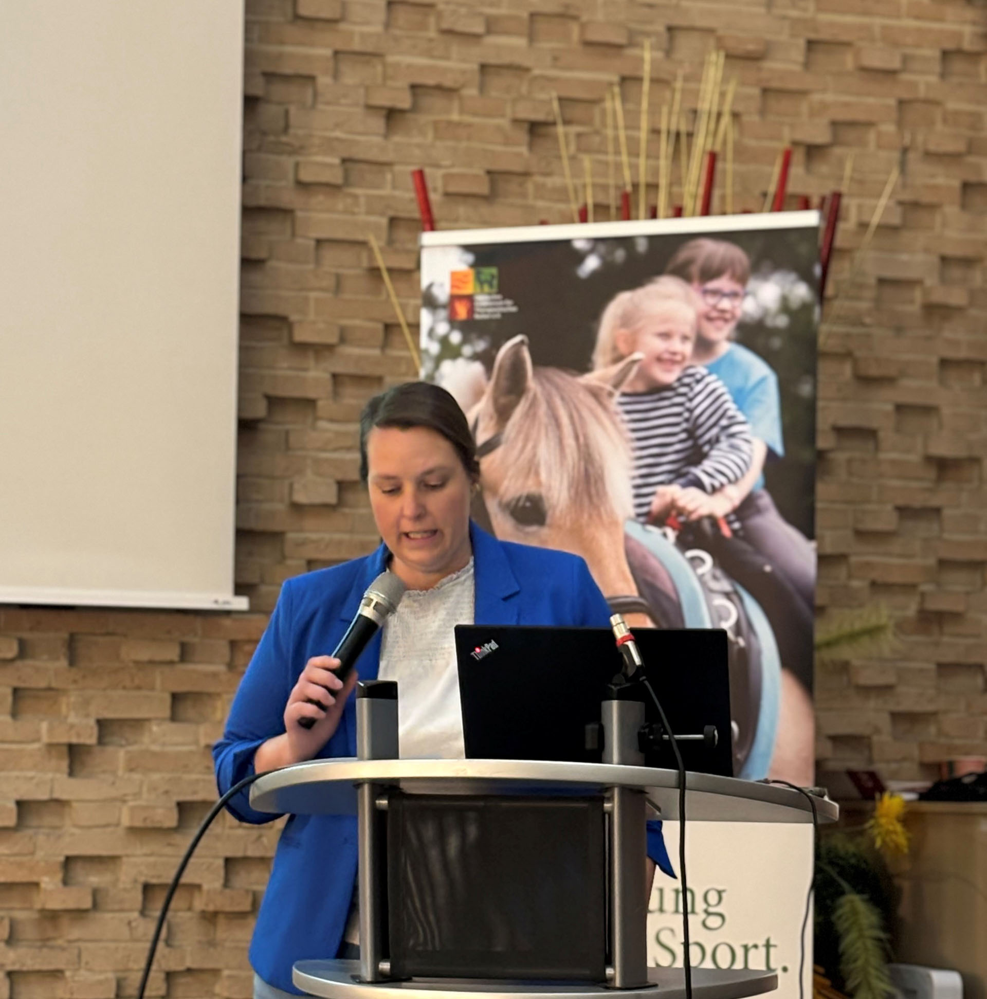 Eine Frau mit einem Mikrofon spricht an einem Podium während einer Präsentation. Sie trägt einen blauen Blazer und schaut auf einen Laptop, während im Hintergrund ein Plakat mit Kindern, die auf einem Pferd sitzen, zu sehen ist. Die Wand ist mit einem strukturierten Steinmuster verziert.