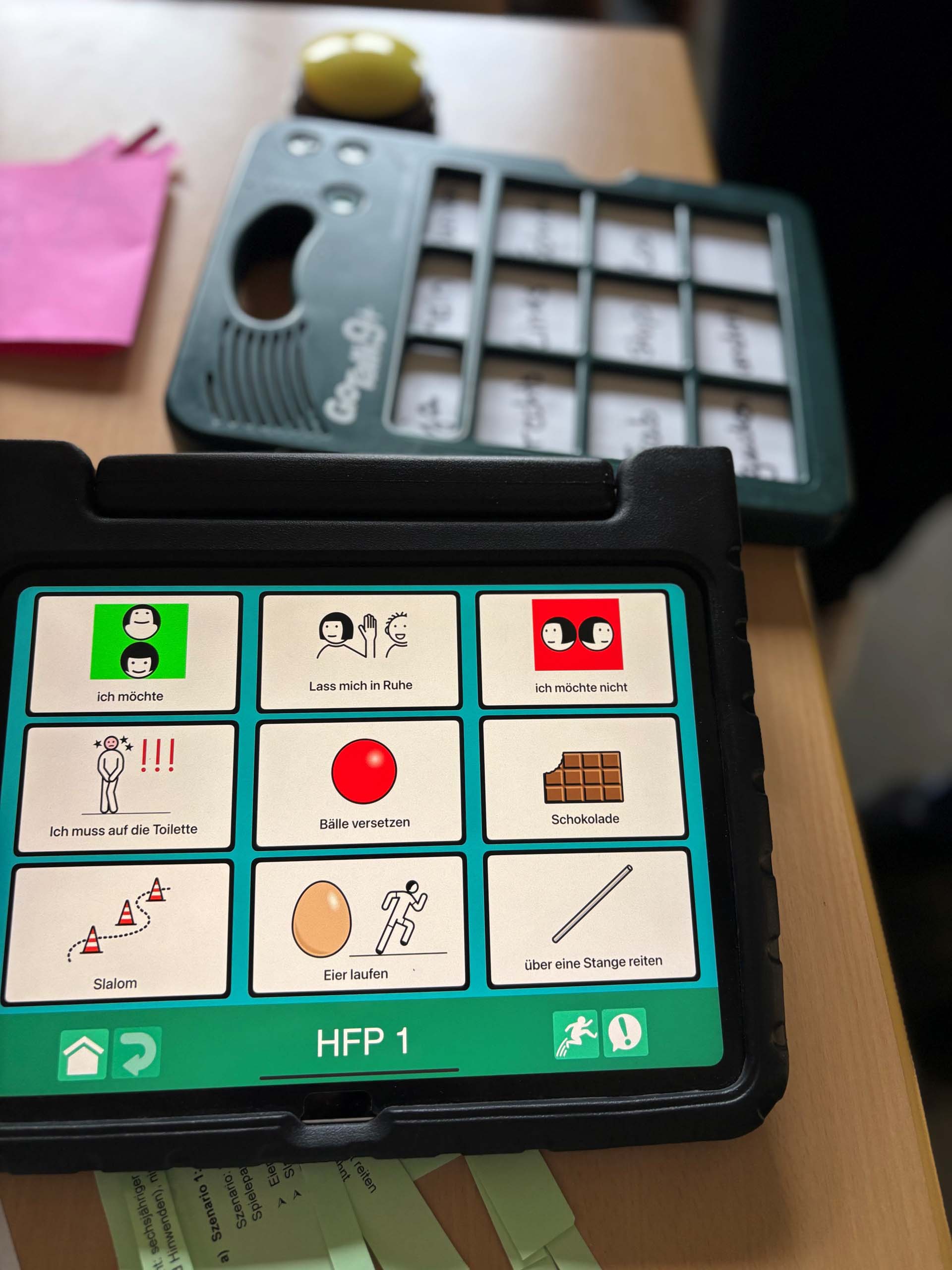 Ein elektronisches Kommunikationsgerät mit Symbolen auf dem Bildschirm, das verschiedene Ausdrücke in deutscher Sprache anzeigt, zusammen mit einem weiteren Gerät im Hintergrund, das eine Reihe von beschrifteten Tasten enthält, auf einem Tisch liegend.