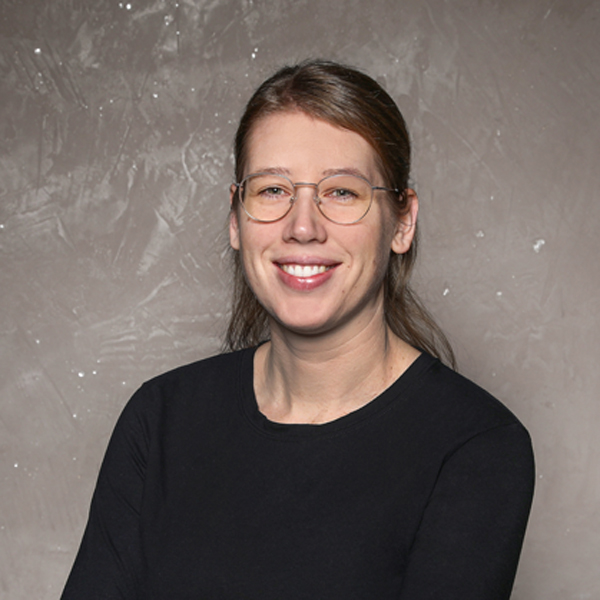 Eine Frau mit Brille und langen, braunen Haaren, die in einem schwarzen Oberteil lächelt, vor einer grauen Wand mit Struktur.