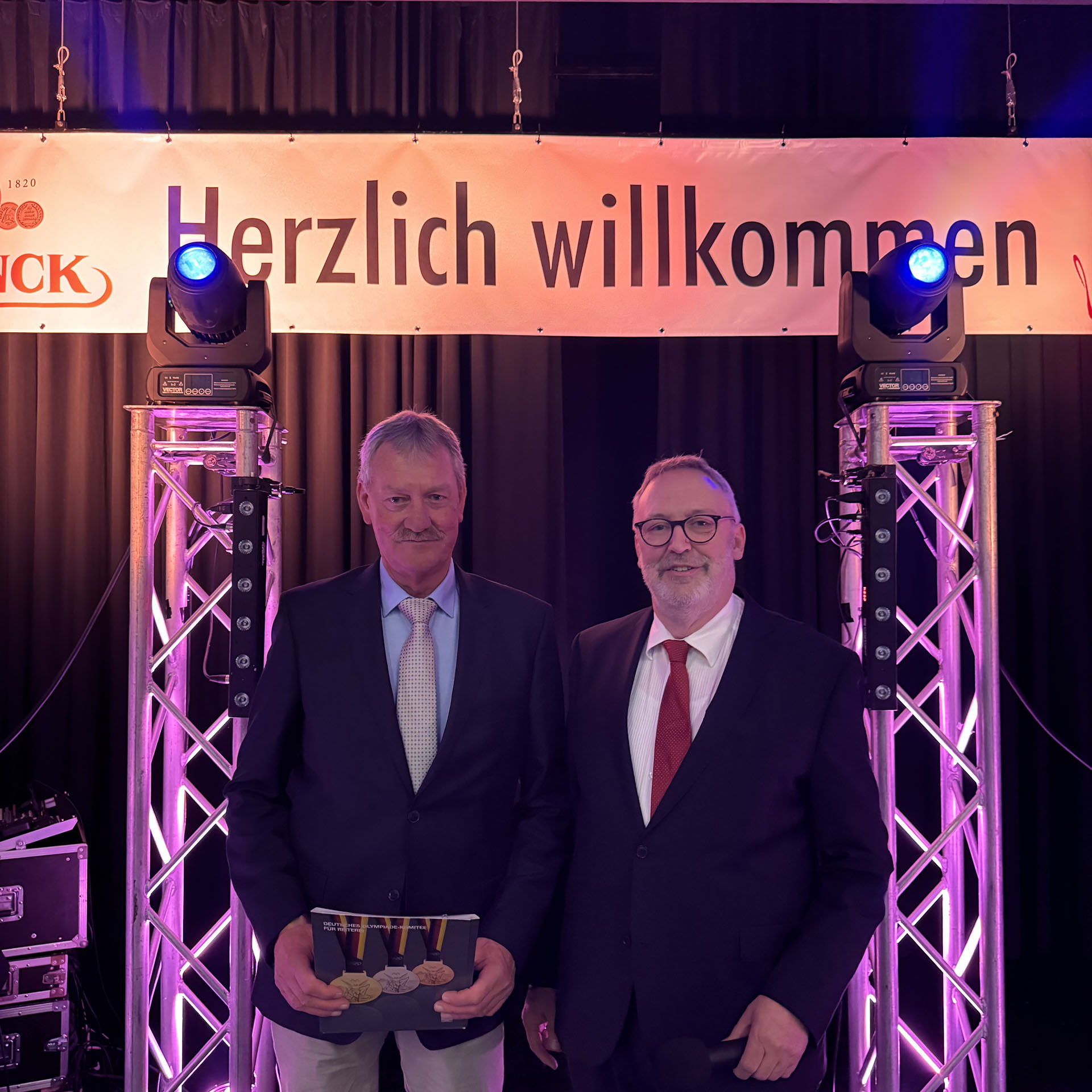 Zwei Männer stehen auf einer Bühne unter einem "Herzlich willkommen"-Schild. Der links trägt Anzug und hat Medaillen in der Hand.